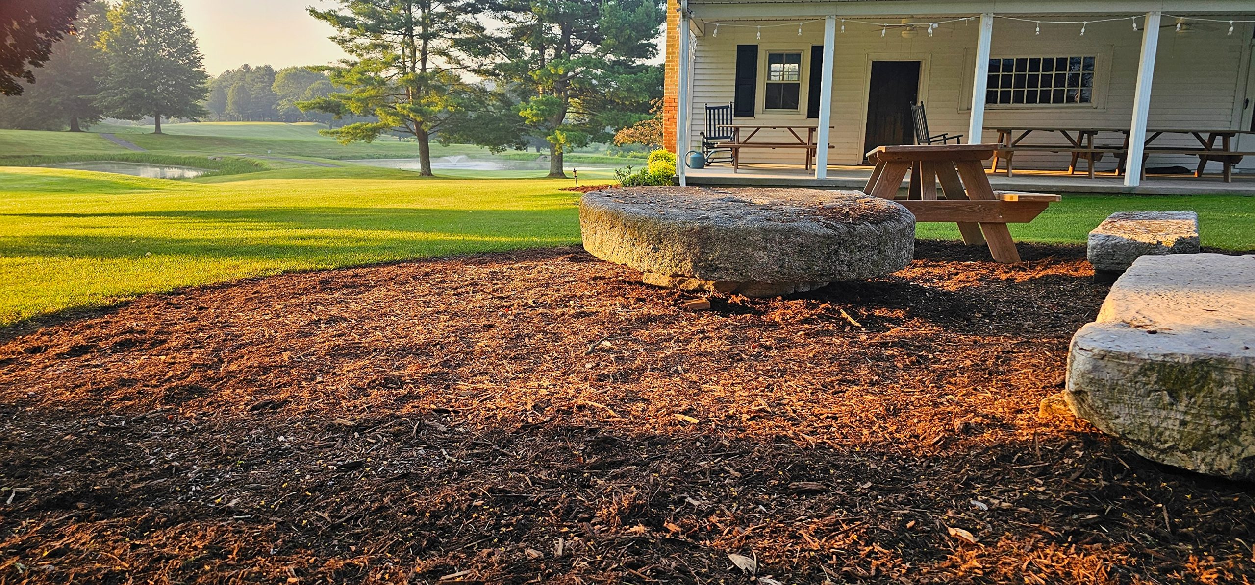 blackberry-hardwood mulch-seating-area-golf-course-landscaping Premium hardwood mulch around a stone seating area near a clubhouse on a manicured golf course at sunrise.