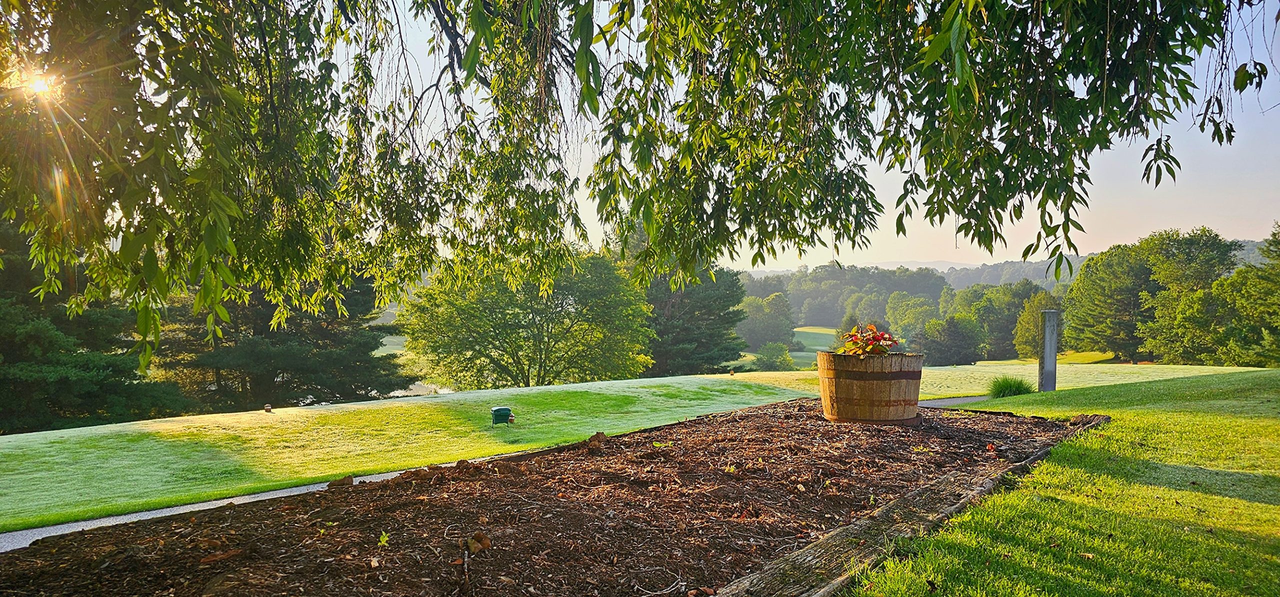 landscape-hardwood-mulch-bed Outdoor garden bed featuring dark premium hardwood mulch and a decorative wooden barrel planter, overlooking a lush green landscape with trees.